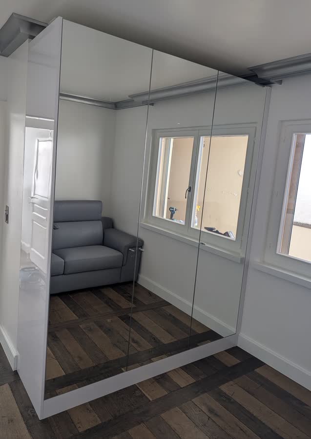 Armoire toute hauteur à façades miroir et flanc laqué blanc, installée dans une pièce claire au parquet foncé.
