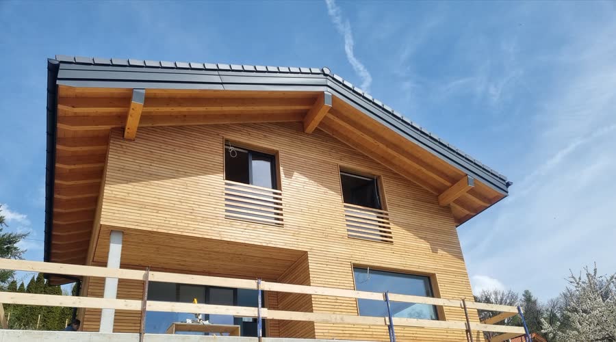 Chalet contemporain en ossature bois avec bardage en lames claires et large avant-toit, sous un ciel bleu.