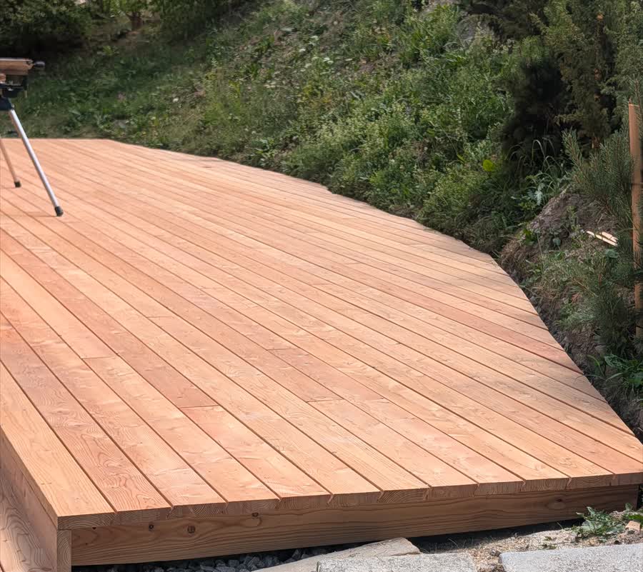 Terrasse en lames de bois neuves, au bord arrondi, posée en bordure d'un talus végétalisé.